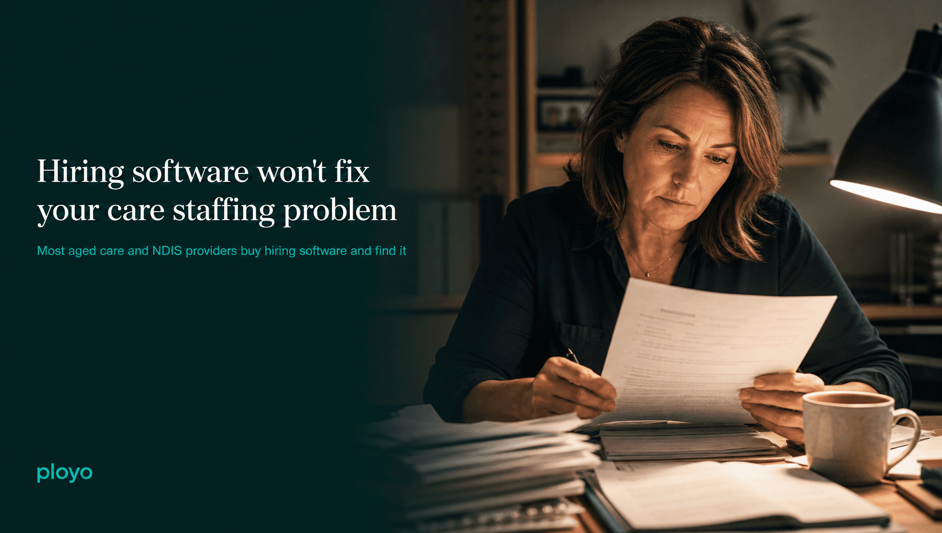 A hiring manager at a desk late in the evening, reviewing printed candidate paperwork under a desk lamp, a cooling cup of coffee within reach