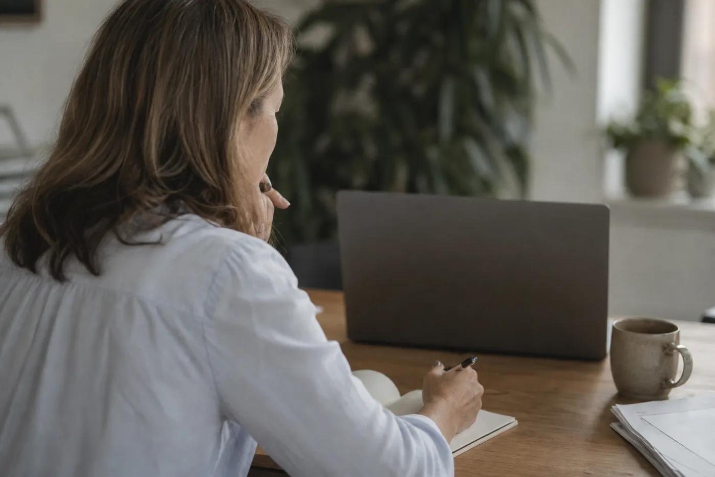 A recruiter at her desk watching a candidate interview back on a laptop, pen in hand