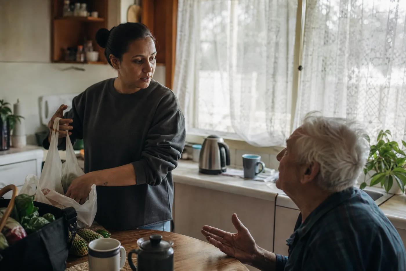A home-care worker in a client's kitchen, talking with him while putting the shopping away